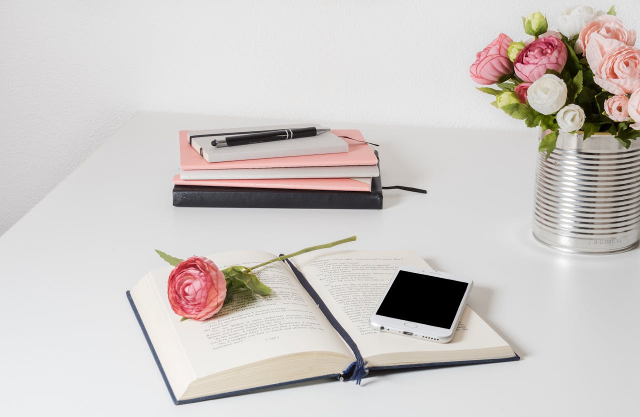 A stylish home office setup with open book, smartphone, and flowers on a white desk.